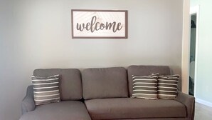 Un sofá seccional gris con almohadones a rayas, un letrero que dice 'welcome' arriba y un piso de madera.