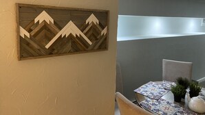Een eetgedeelte met een tafel gedekt voor twee personen, een decoratief houten object aan de muur en een stoel met kussen.