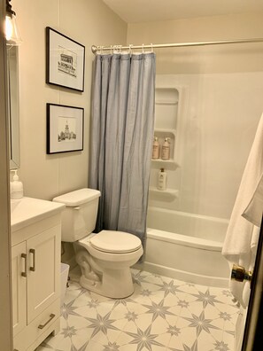 Une salle de bain équipée de toilettes, d’un lavabo, d’une douche et d’une baignoire. Des tableaux encadrés ornent le mur et une serviette est accrochée à la porte.