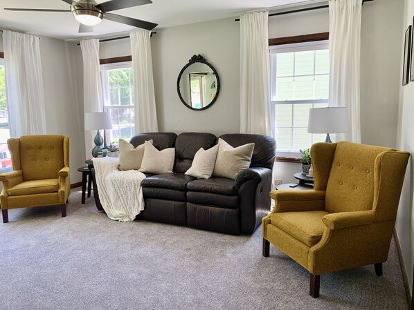 Un salon avec un canapé en cuir foncé, deux fauteuils de couleur moutarde, un ventilateur de plafond et un grand miroir au mur.