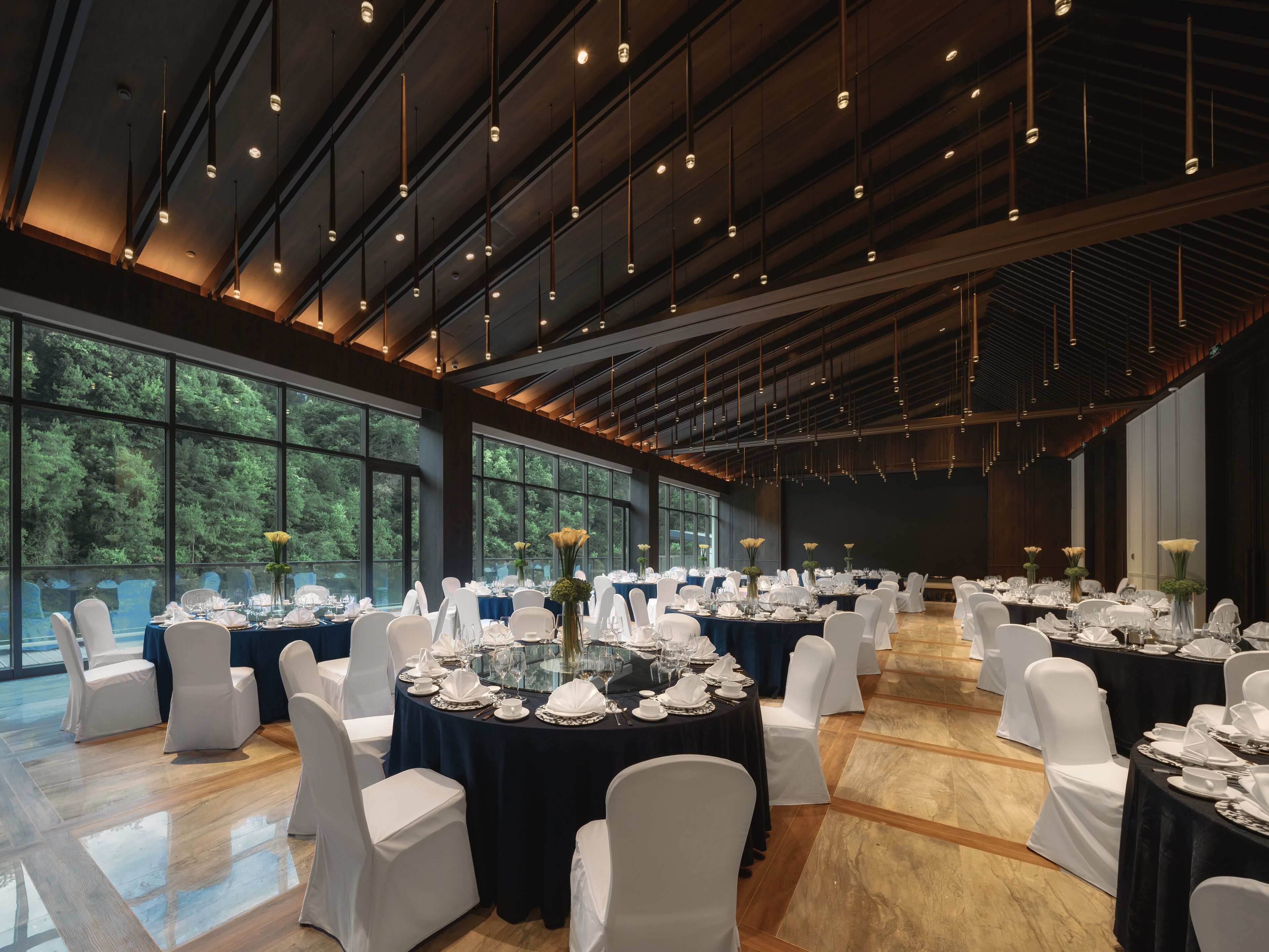Un gran salón de banquetes con mesas redondas dispuestas para comer, sillas blancas y vistas a árboles a través de amplias ventanas.