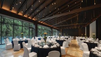 Un gran salón de banquetes con mesas redondas dispuestas para comer, sillas blancas y vistas a árboles a través de amplias ventanas.