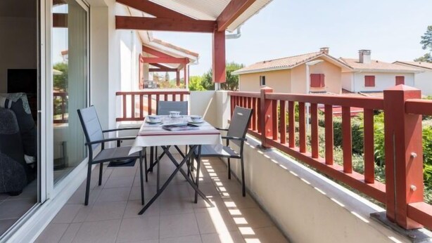 A balcony with a table and chairs, a red railing, and a view of neighboring houses. - Côté du Lac by Interhome (Soustons)