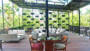 Terraza al aire libre con mobiliario de mimbre, una mesa con superficie de vidrio y un fondo de macetas verdes.