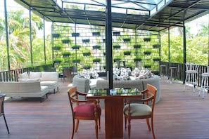 Terraza al aire libre con mobiliario de mimbre, una mesa con superficie de vidrio y un fondo de macetas verdes.