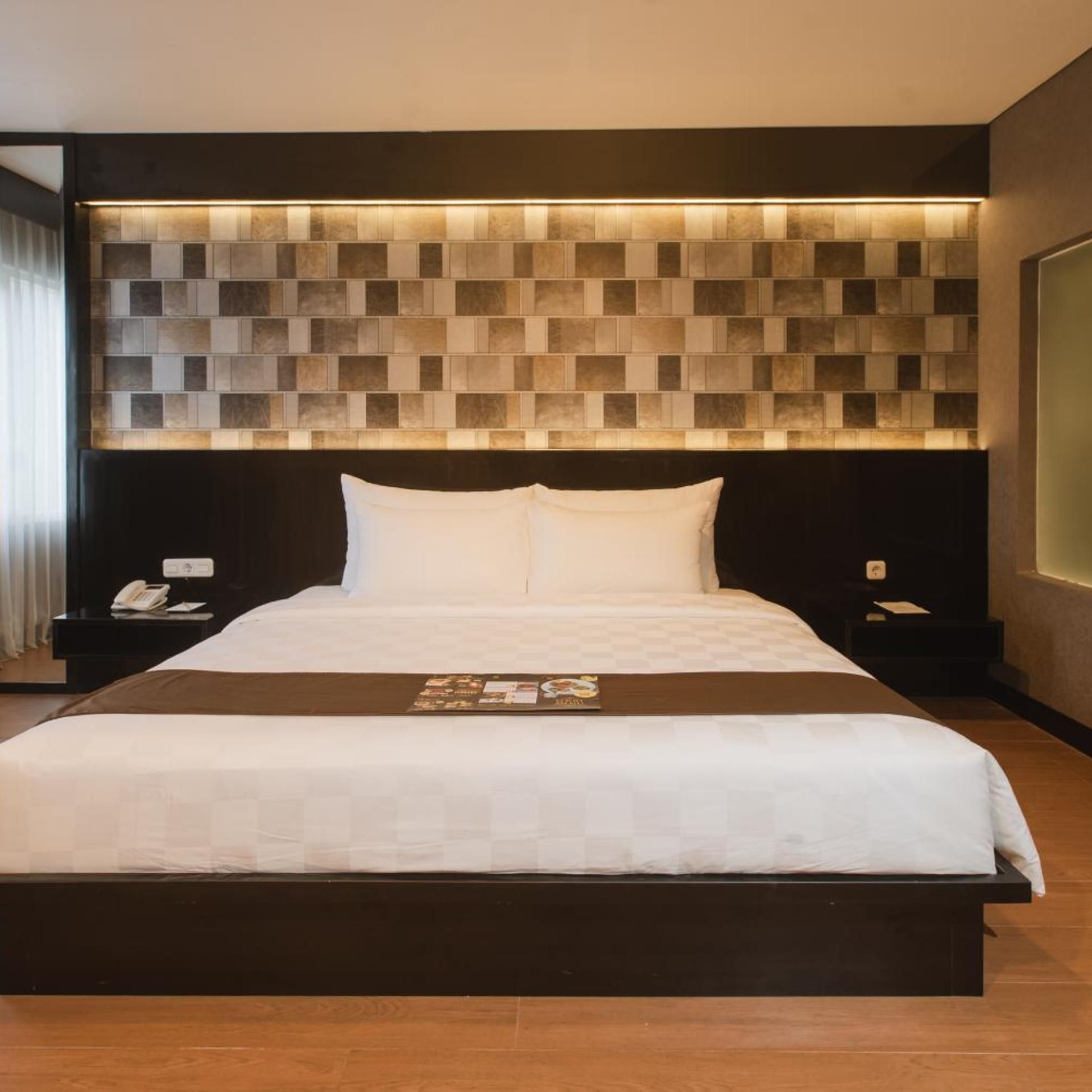 A modern hotel room with a large bed, a headboard with a geometric tile design, a bedside table with a phone, and a nightstand with a lamp.