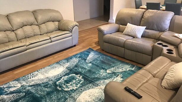 Een woonkamer met twee beige leren banken, een vloerkleed met blauw patroon, en een afstandsbediening op een van de banken.
