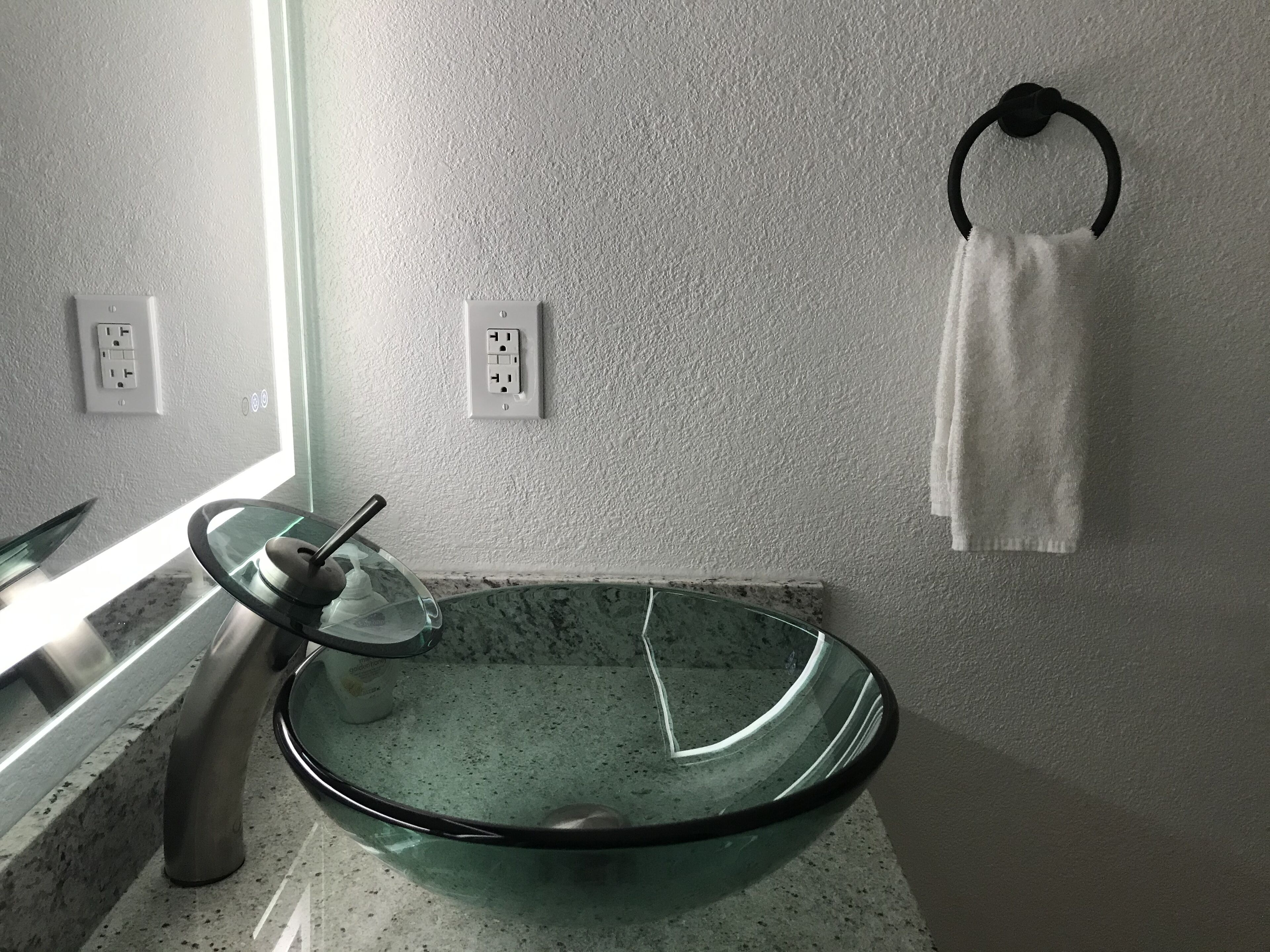 Un baño con un lavamanos de cristal, un perchero y dos enchufes en la pared.