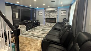 A modern living room with a large flat-screen TV, a fireplace, a black leather sofa, and a patterned rug. - Cape Cod Sunscape. Orleans (Orleans)