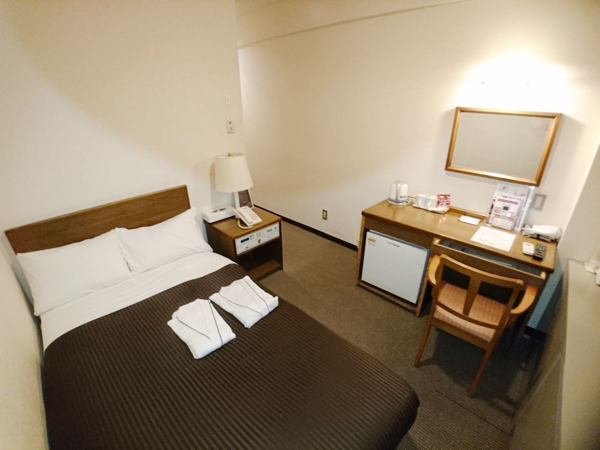 Ein Hotelzimmer mit Bett, Schreibtisch, Stuhl, Nachttisch, Lampe, Telefon, Spiegel und kleinem Kühlschrank.