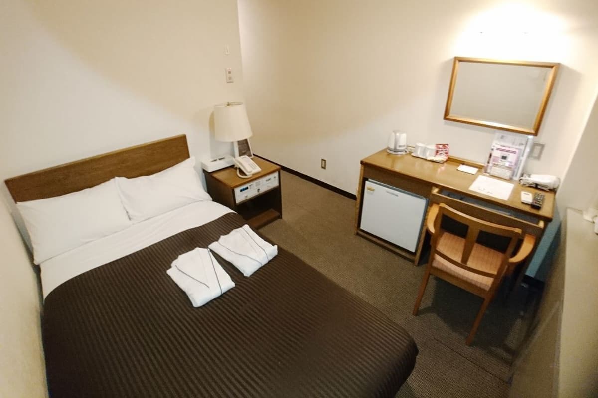 A hotel room with a bed, a desk, a chair, a nightstand, a lamp, a phone, a mirror, and a small refrigerator.