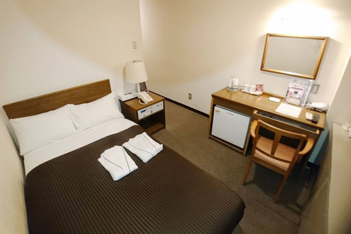 Ein Hotelzimmer mit Bett, Schreibtisch, Stuhl, Nachttisch, Lampe, Telefon, Spiegel und kleinem Kühlschrank.