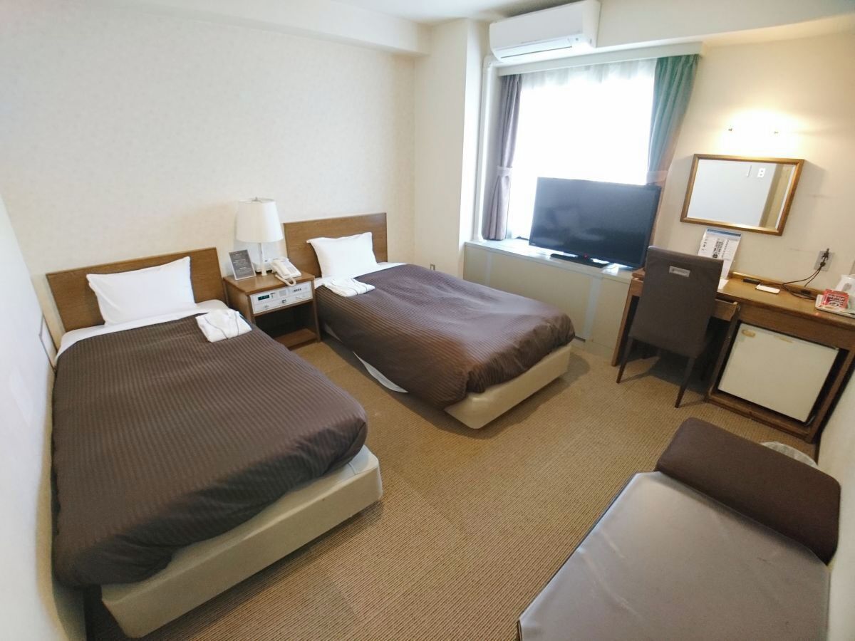 Ein Hotelzimmer mit zwei Betten, einem Schreibtisch, einem Stuhl, einem Fernseher und einem kleinen Kühlschrank.