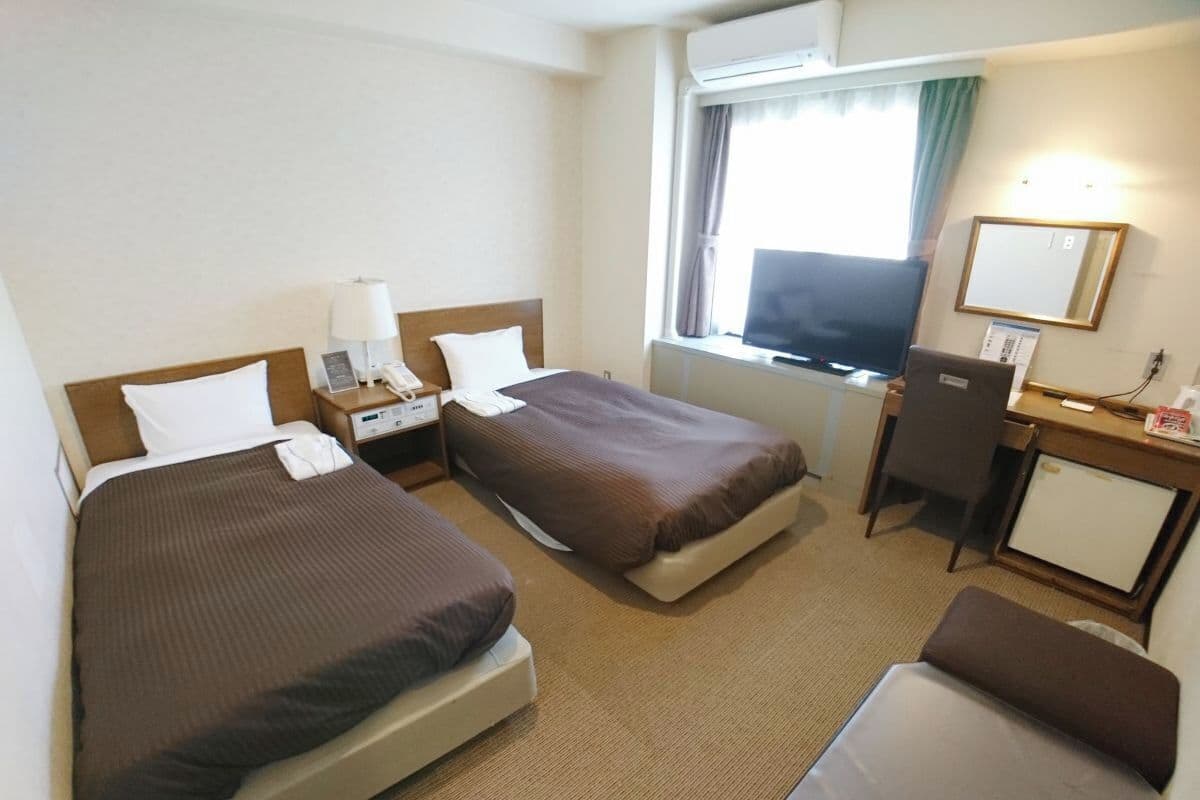 A hotel room with two beds, a desk, a chair, a TV, and a small refrigerator.