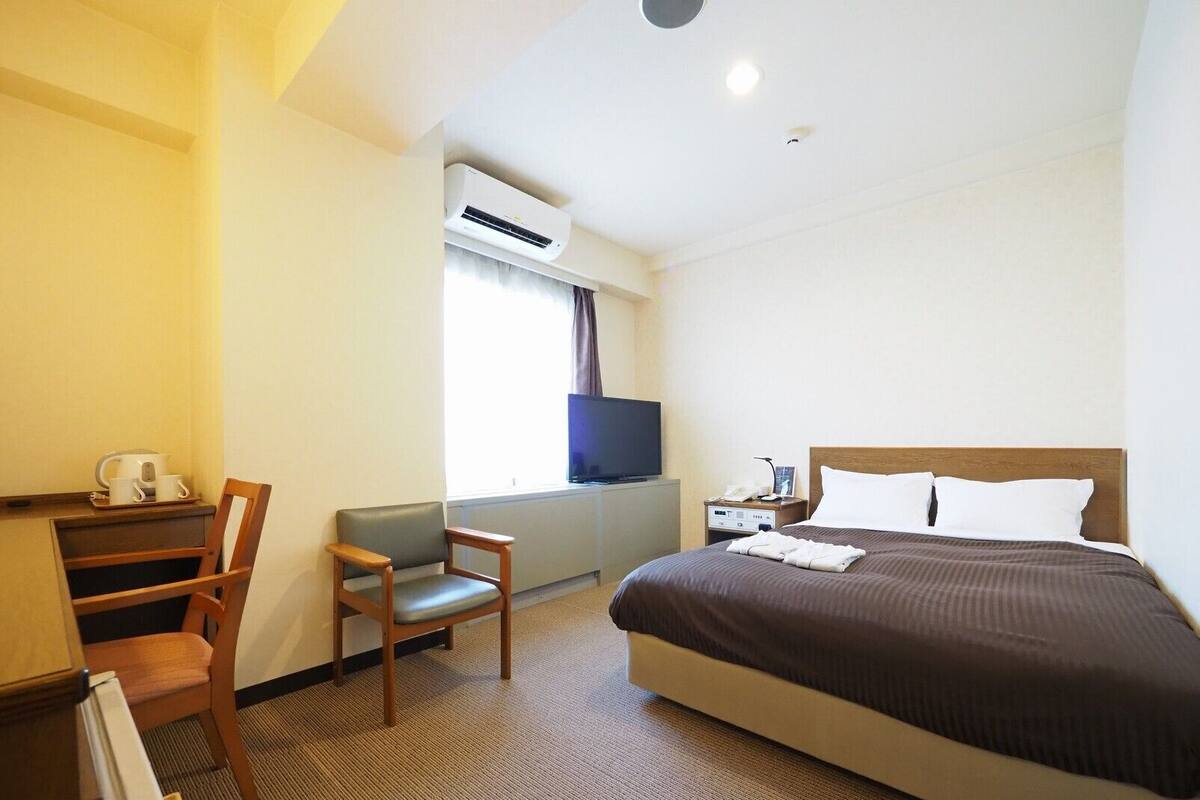 A hotel room with a bed, a desk with a chair, a television, and a window with curtains.