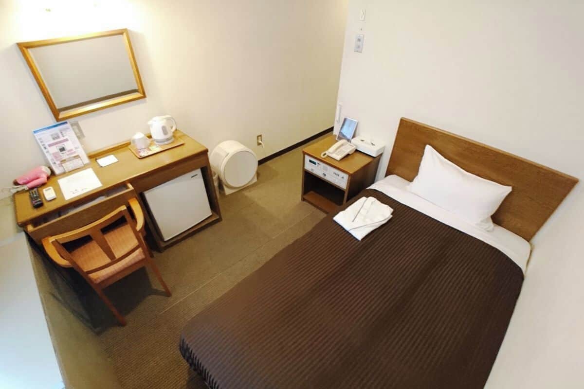 A hotel room with a bed, desk, chair, and a small refrigerator.