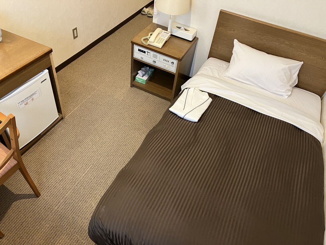 Ein Hotelzimmer mit einem Bett, einem Nachttisch mit Telefon und Lampe, einem Schreibtisch mit Kühlschrank und einem Sessel.