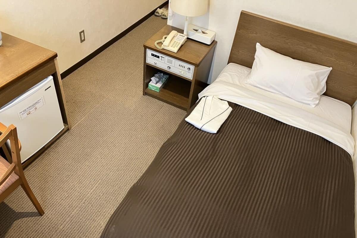 A hotel room with a bed, a nightstand with a phone and lamp, a desk with a refrigerator, and a chair.