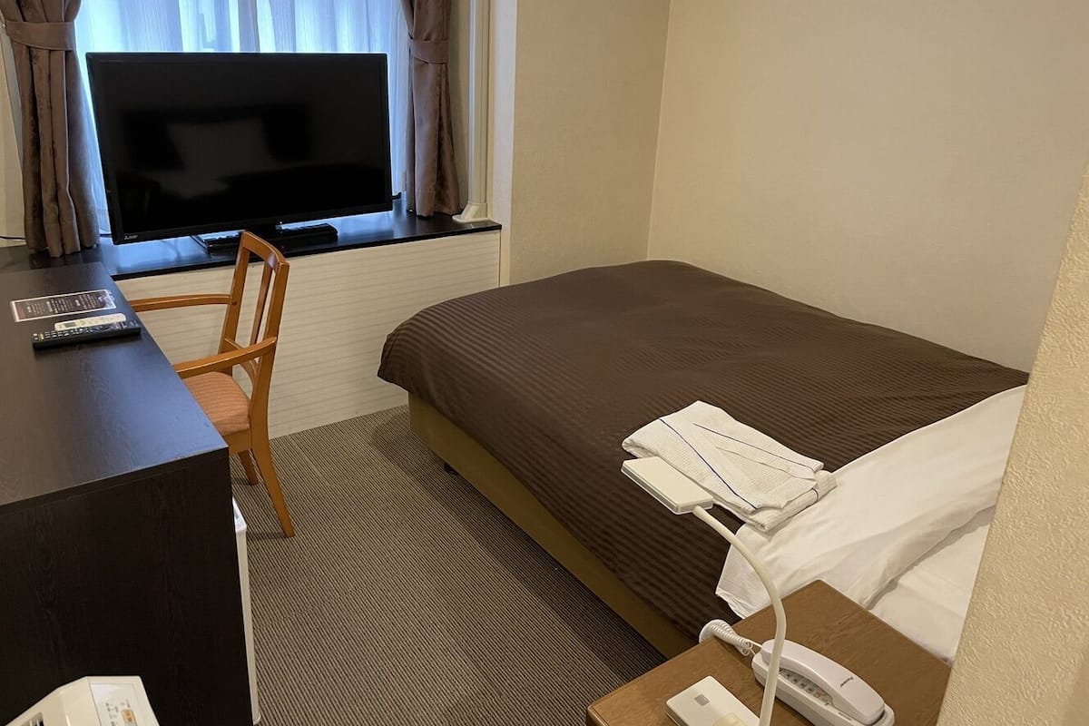 A hotel room with a bed, a desk, a chair, a TV, and a window with curtains.