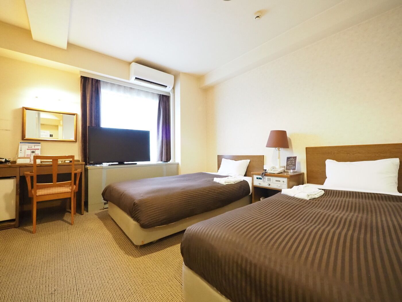 Ein Hotelzimmer mit zwei Betten, einem Schreibtisch, einem Stuhl, einer Klimaanlage und einem Fenster mit Vorhängen.