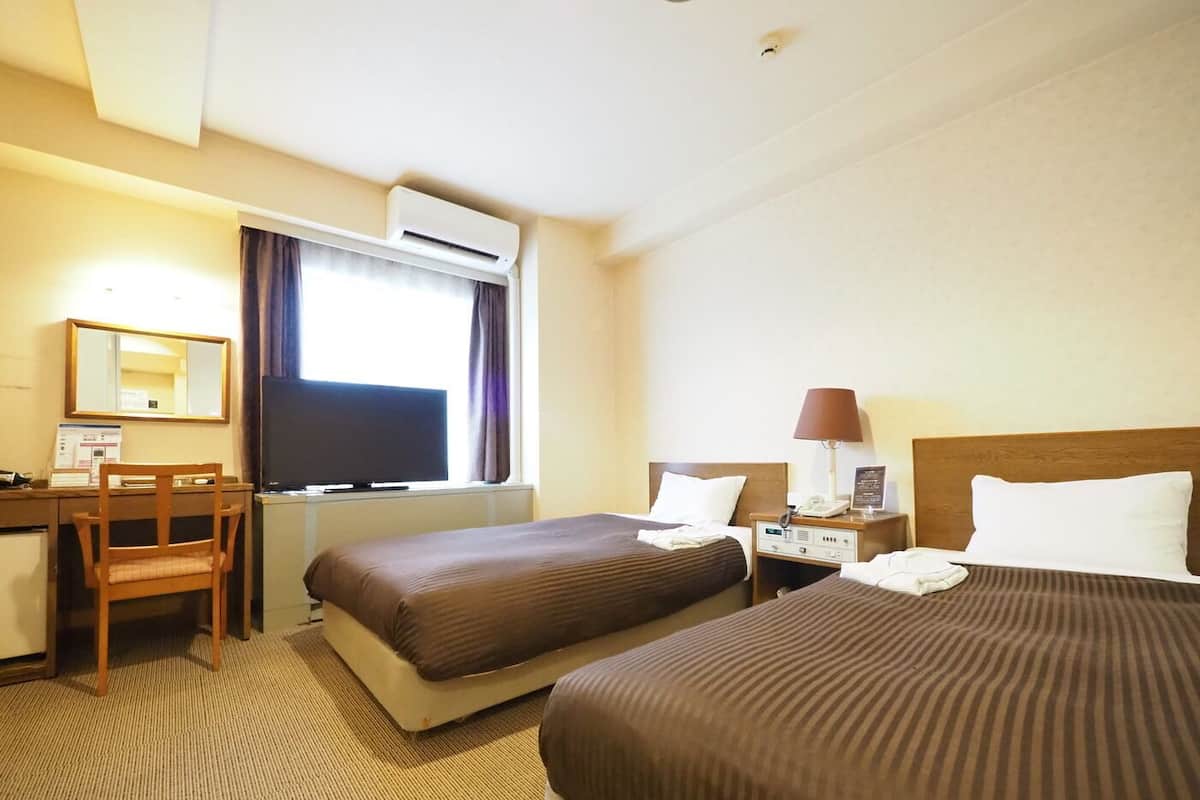 A hotel room with two beds, a desk, a chair, an air conditioning unit, and a window with curtains.