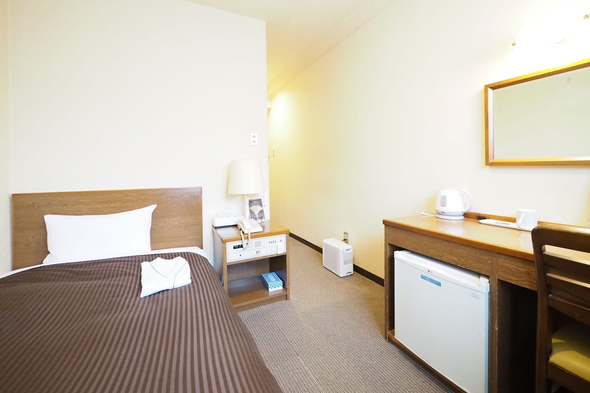 A hotel room with a bed, bedside table, lamp, desk, chair, and a small refrigerator.
