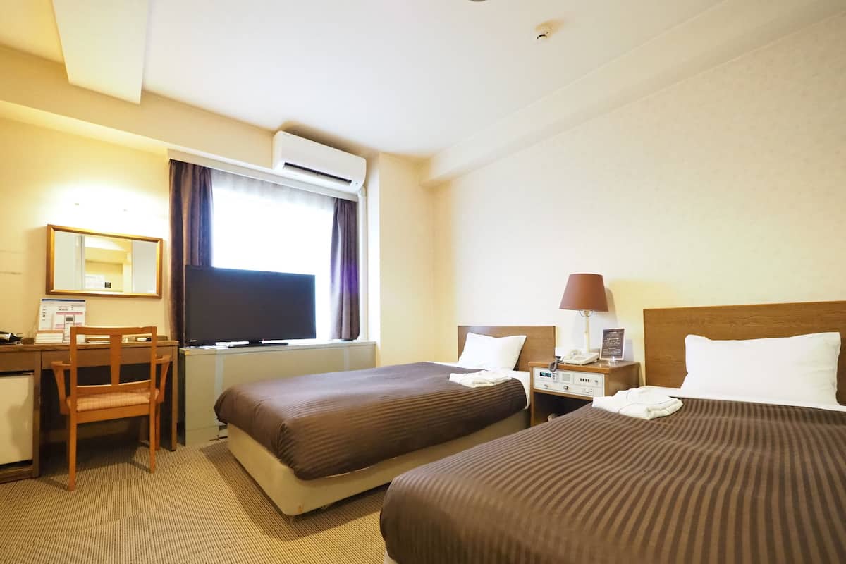 Ein Hotelzimmer mit zwei Betten, einem Schreibtisch, einem Stuhl, einer Klimaanlage und einem Fenster mit Vorhängen.