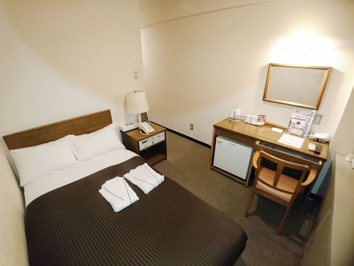 Ein Hotelzimmer mit Bett, Schreibtisch, Stuhl, Nachttisch, Lampe, Kühlschrank und Spiegel.