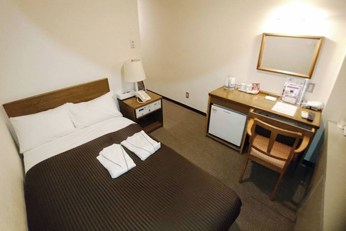A hotel room with a bed, a desk, a chair, a nightstand, a lamp, a refrigerator, and a mirror.
