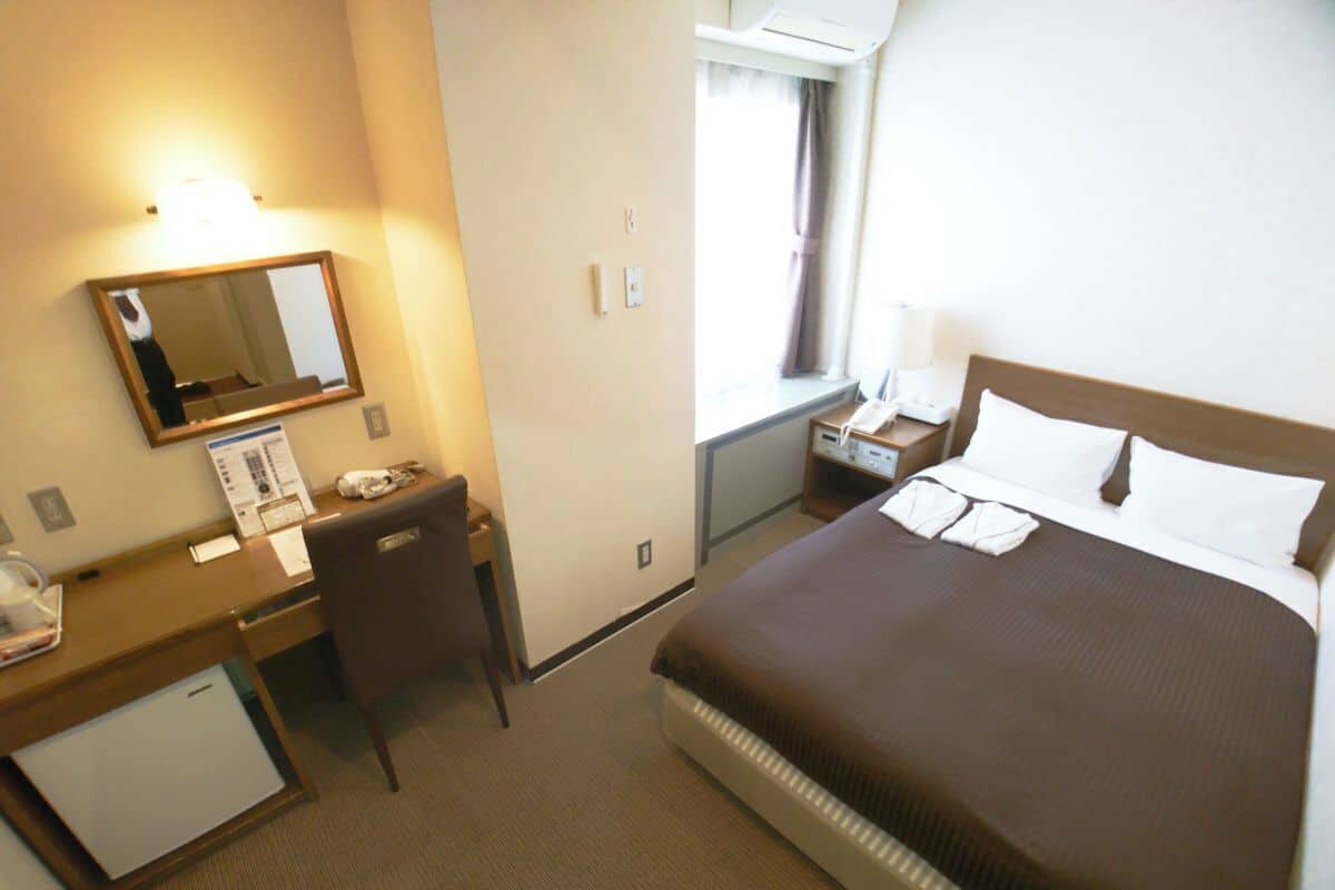 A hotel room with a bed, desk, chair, and a small refrigerator.