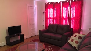 Una sala de estar con un sofá, una mesa de centro de vidrio, un televisor sobre un mueble y cortinas rojas.