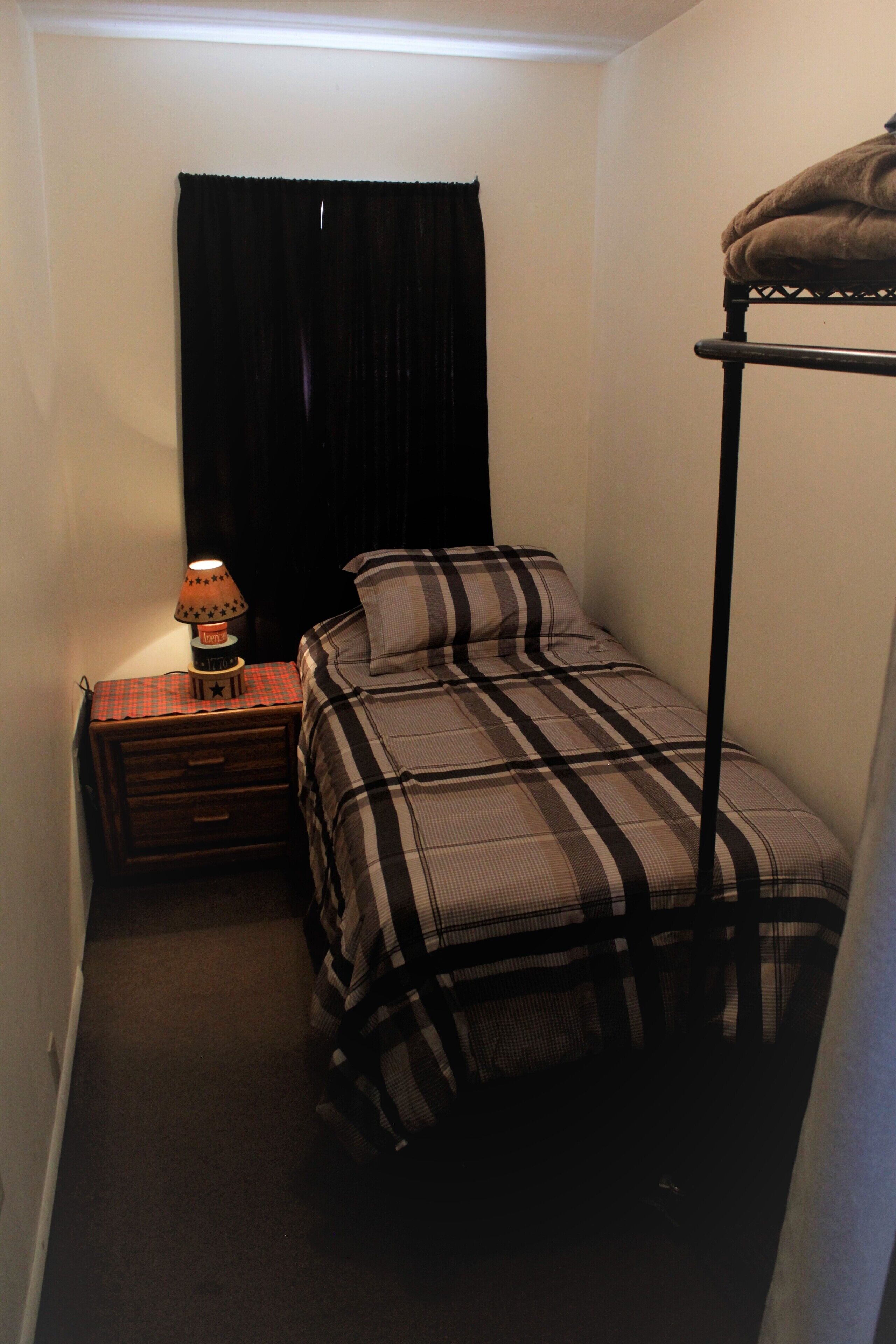A small, dimly lit room with a single bed, a nightstand with a lamp, and a window with black curtains.