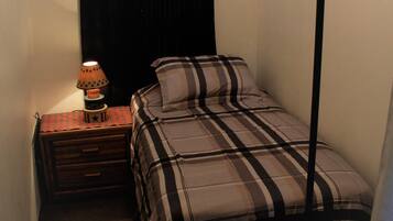A small, dimly lit room with a single bed, a nightstand with a lamp, and a window with black curtains.