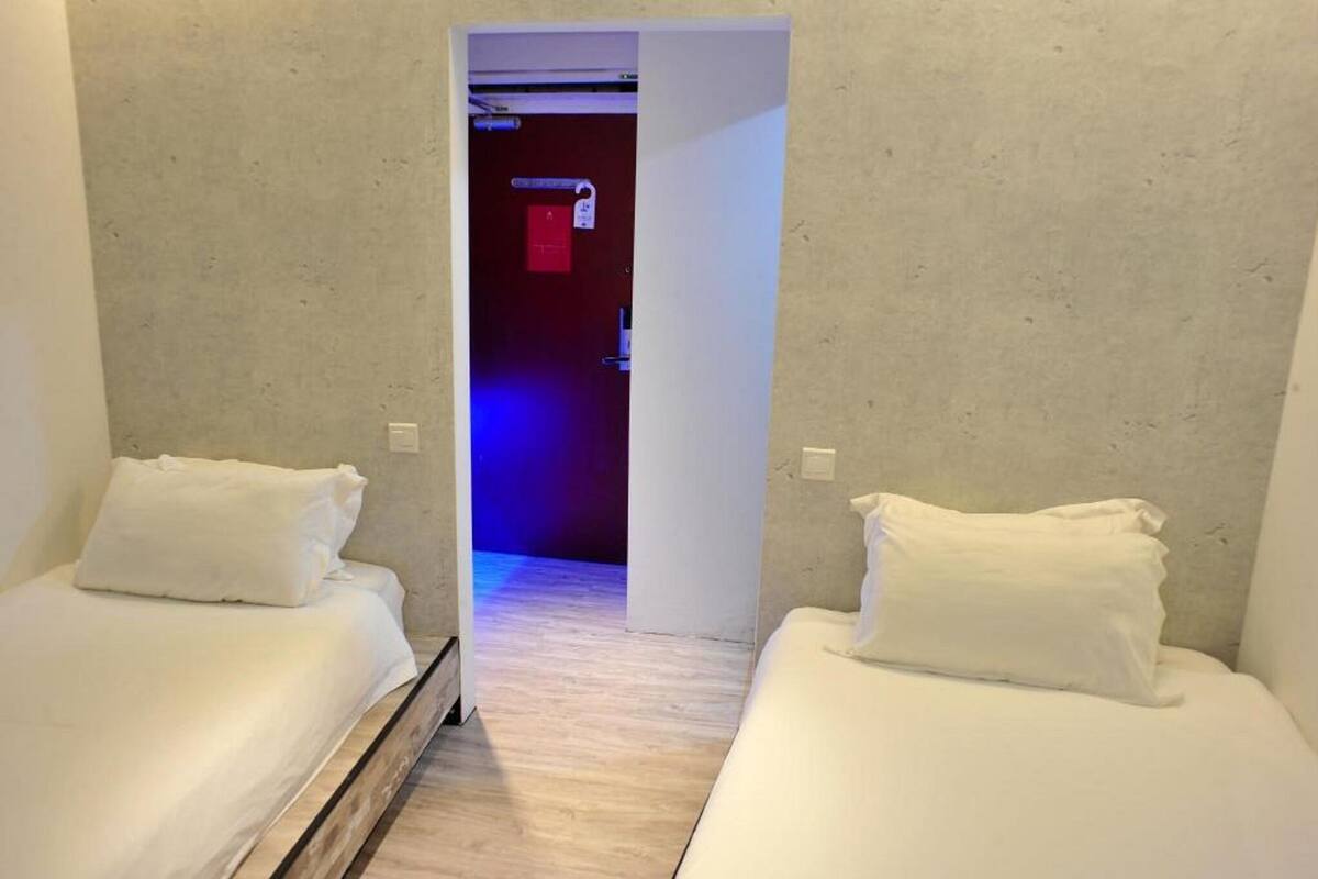 A hotel room with two single beds, a wooden floor, and a hallway leading to a bathroom with a red door.