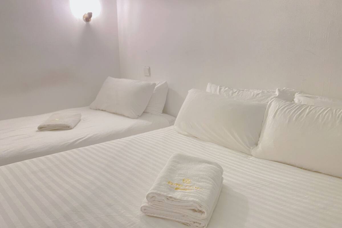 A hotel room with two beds, each with white linens and pillows, a folded towel on the floor, and a wall-mounted light fixture.