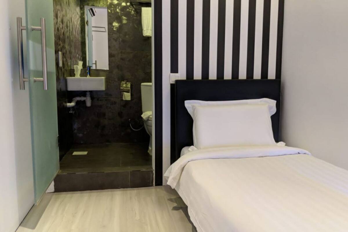 A small, modern room with a bed, a bedside table, a mirror, and a bathroom visible through an open door.