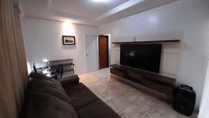A living room with a large flat-screen TV mounted on the wall, a brown sofa, a desk with a chair, and a suitcase on the floor. - Exclusive Pool  (Planaltina)