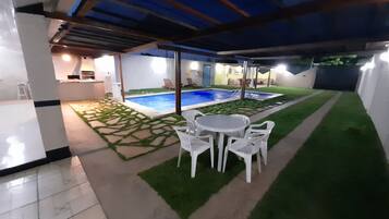Un espacio al aire libre techado con piscina, sillas de plástico blancas y una mesita.