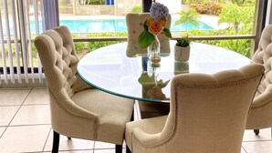 Une table ronde en verre, entourée de quatre chaises rembourrées, un vase rempli de fleurs et une vue sur une piscine et la verdure environnante.