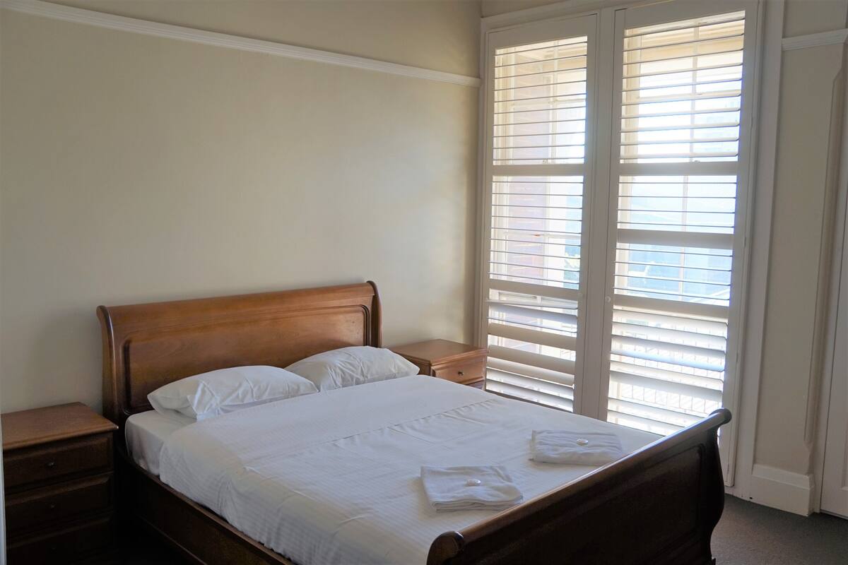 A neatly made bed with white linens and two pillows, a wooden headboard, a nightstand with a drawer, and a window with blinds.