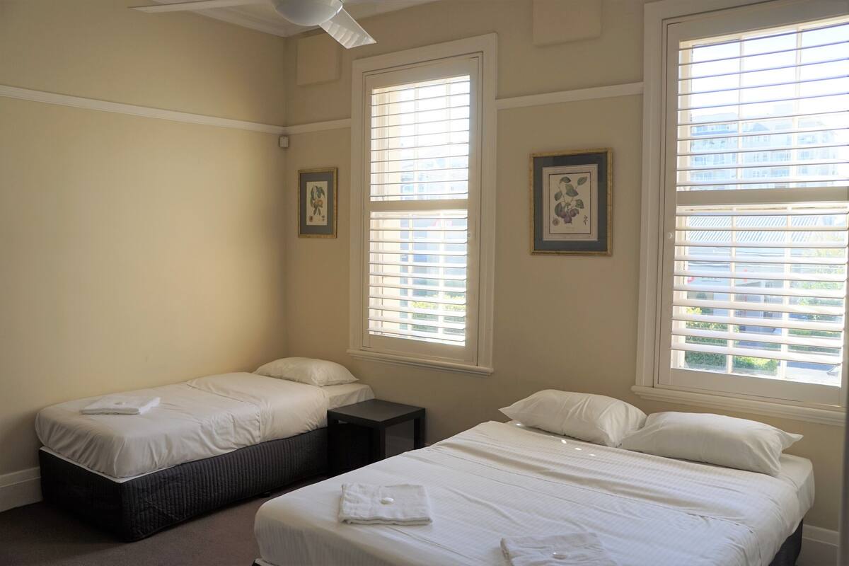 A bedroom with two single beds, a ceiling fan, and large windows with blinds.