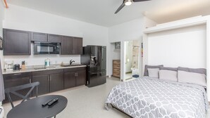 A compact hotel room with a kitchenette, a bed with patterned bedding, a ceiling fan, and a small table with a remote control. - Chugach Mountains View South Anch Premium Suite (Anchorage)