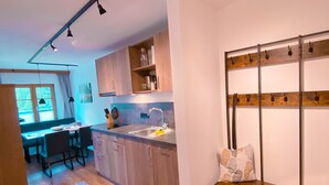 Een moderne keuken met houten kastjes, een ingebouwde gootsteen en hanglampen. Er is een zithoek met een tafel en stoelen, en een bank met haken aan de muur.