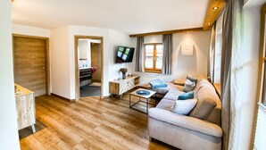 Ein modernes Wohnzimmer mit Sofa, Couchtisch und Fernseher. Ein Holzboden und eine Tür zum Badezimmer sind vorhanden.