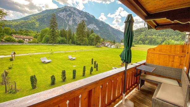 Ein hölzerner Balkon mit Blick auf eine grüne Wiese, einen Berg und ein Haus im Hintergrund.