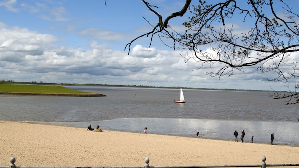 Een zandstrand met een rustig wateroppervlak, een zeilboot en mensen aan de kust.