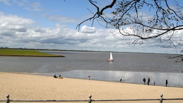 Une plage de sable fin, une étendue d’eau calme, un voilier et des personnes se trouvant au bord de l’eau.