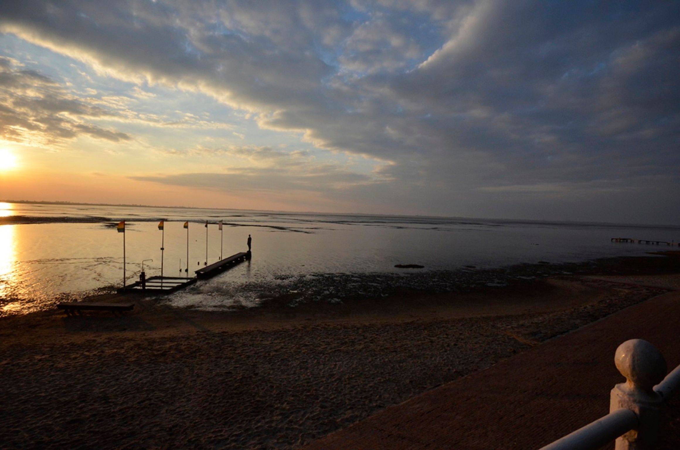 Une plage avec une jetée qui s’avance dans des eaux calmes, un banc et une statue.