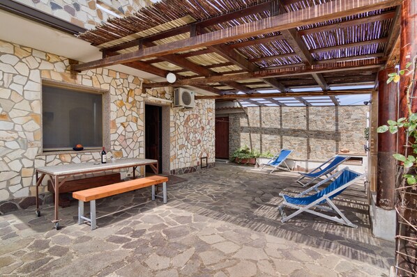 Un patio de piedra con mesa y banco de madera, dos sillones azules y una pérgola.