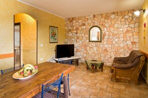 A dining area with a wooden table, chairs, a television, and a stone wall. - Appartamento Nuraghe with Air Conditioning, Garden and Terrace (Gonnesa)